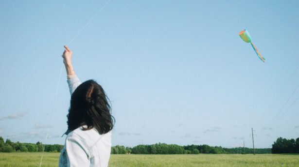 person with dark hair flying a kite in an open field against a blue sky